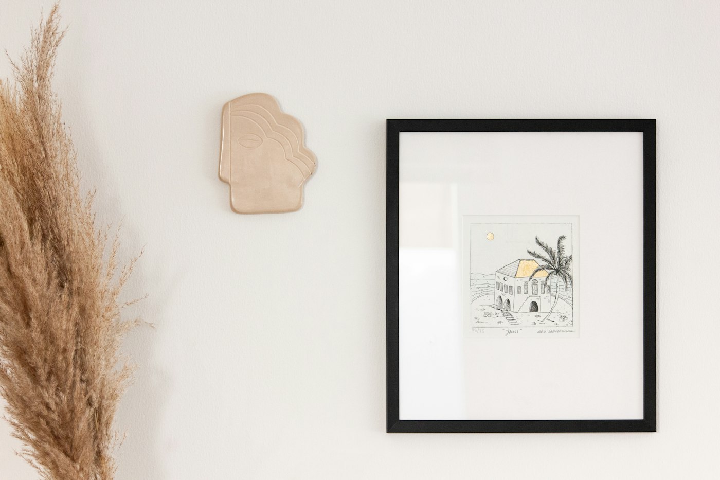 A framed print on a white wall beside dried pampas grass
